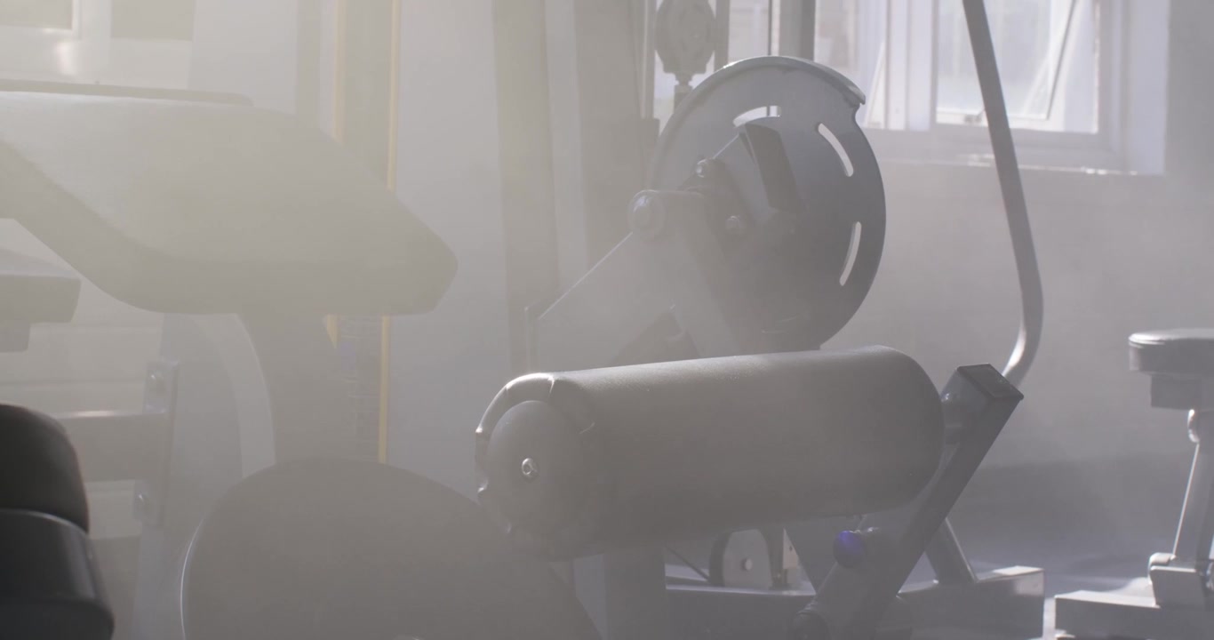 Camera is panning as window light is clearing haze, showing leg curl machine at gym