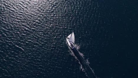 Velero-Blanco-Navegando-Por-El-Mar-Azul-Con-Sol-Brillante-En-La-Superficie,-Aéreo