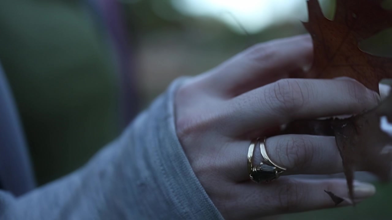 A woman’s hand reaches through autumn leaves to pick up a rolled scroll in soft, natural outdoor light. Intimate close-up footage highlights subtle textures and contemplative mood.