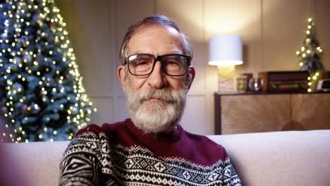 close up of cheerful smiling old male pensioner in glasses chatting on video call in room with glowing decorated xmas tree