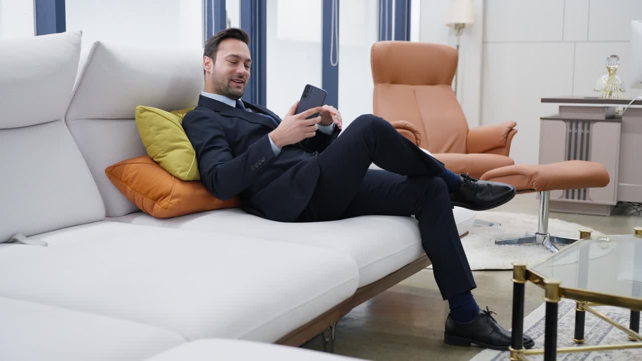 Happy businessman enjoying success win on mobile phone on sofa at the premium office. Joyful male reading good news on smartphone, showing yes gesture, celebrating victory on phone in luxury room