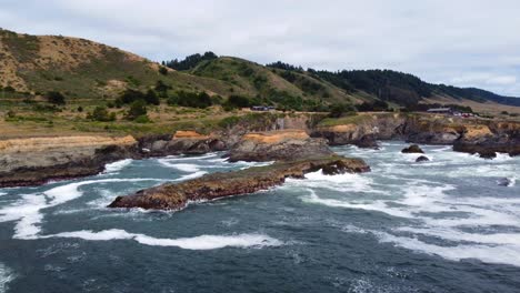 4K-30FPS-Aerial-Footage-Oregon-Coast---Tracking-in-on-a-coastal-mossy-rock-as-the-ocean-waves-crash-against-oceanside-rock-formations-and-seagull-birds-fly-by-PNW-forests-and-epic-homestead-background