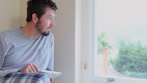 Young-man-looking-out-side-through-window-while-using-tablet-4K-4k