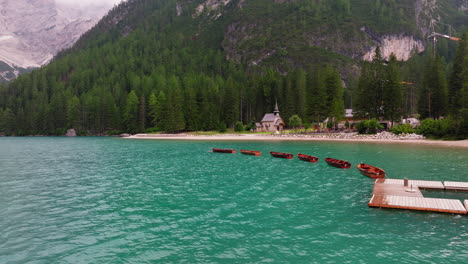 lago di braies or lake braies crystal blue lake with wooden rowing boats in italian dolomites, lago di braies chapel and coniferous and broadleaf trees in background, drone shot