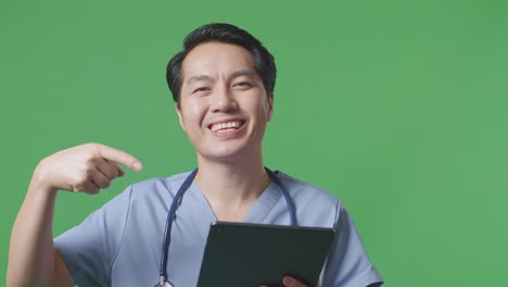 병원 에서 녹색 스크린 배경 에 서 있는 동안 손 에 태블릿 을 가리키는 스테토스코프 를 들고 있는 아시아 남자 의사 의 근접 사진