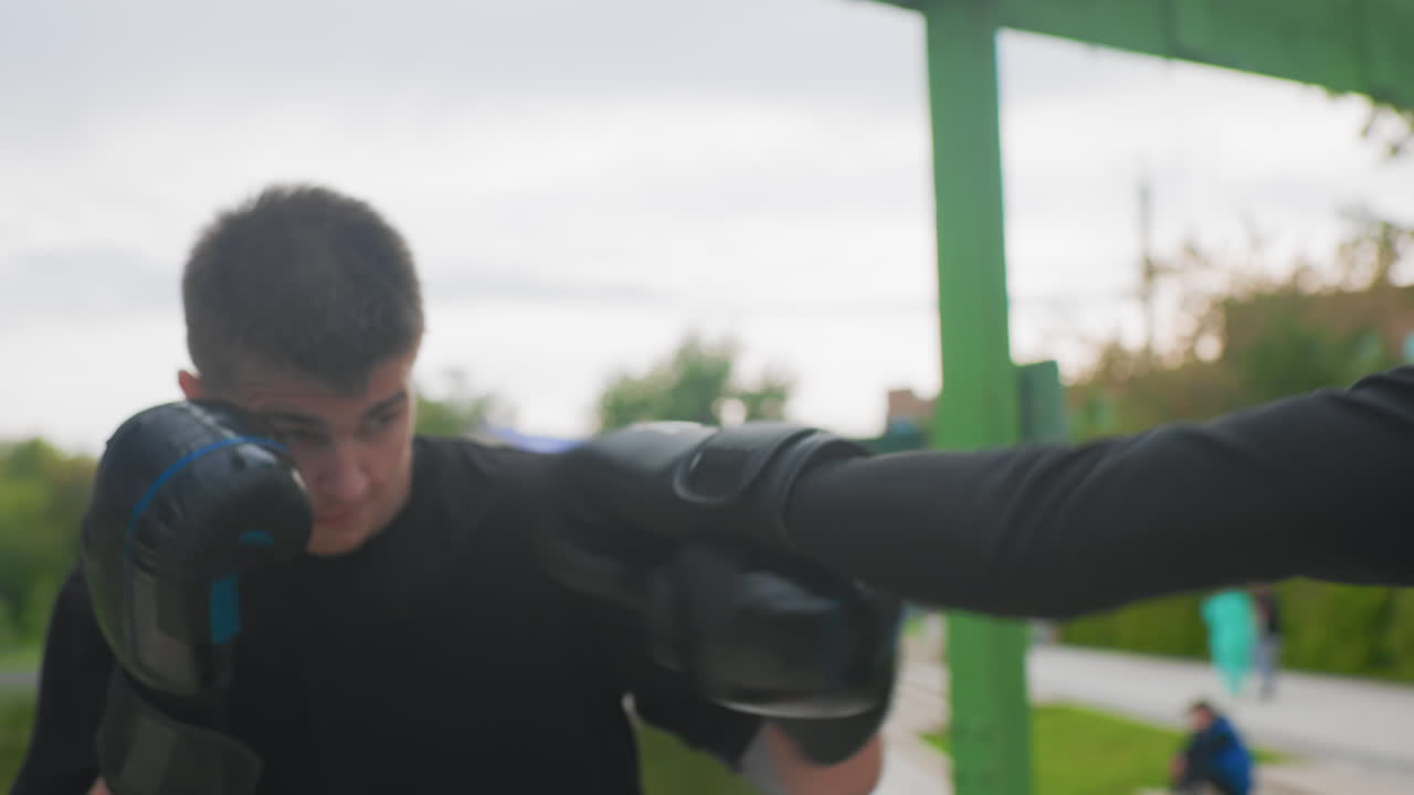 Tense caucasian trainee defends with gloves, highguard protection and tight elbows, focused gaze and quick reflexes, coachguided counter drills on riverside walkway, trees and lake backdrop