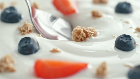 granola with fruit scooped with silver spoon from creamy thick yogurt in macro and slow motion