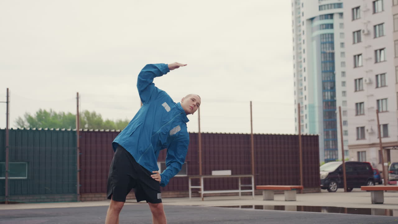 trainer uomo che si riscalda su un campo urbano, esercizi di mobilità con piegamenti laterali, giacca a vento e pantaloncini blu, torri di appartamenti sullo sfondo, canestro da basket vuoto, preparazione mirata per la sessione di allenamento di calcio