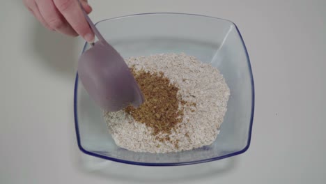 pouring half cup of milled flaxseed on top of rolled oats in glass bowl, closeup