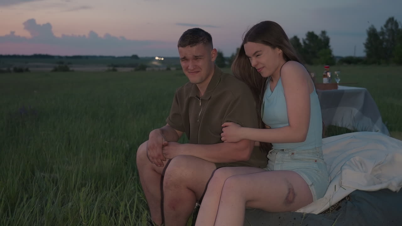 couple sitting on cushioned blanket in open farmland enjoying sunset picnic reacts with laughter and surprise as blades of grass fly around them in gentle breeze, champagne glasses on nearby table