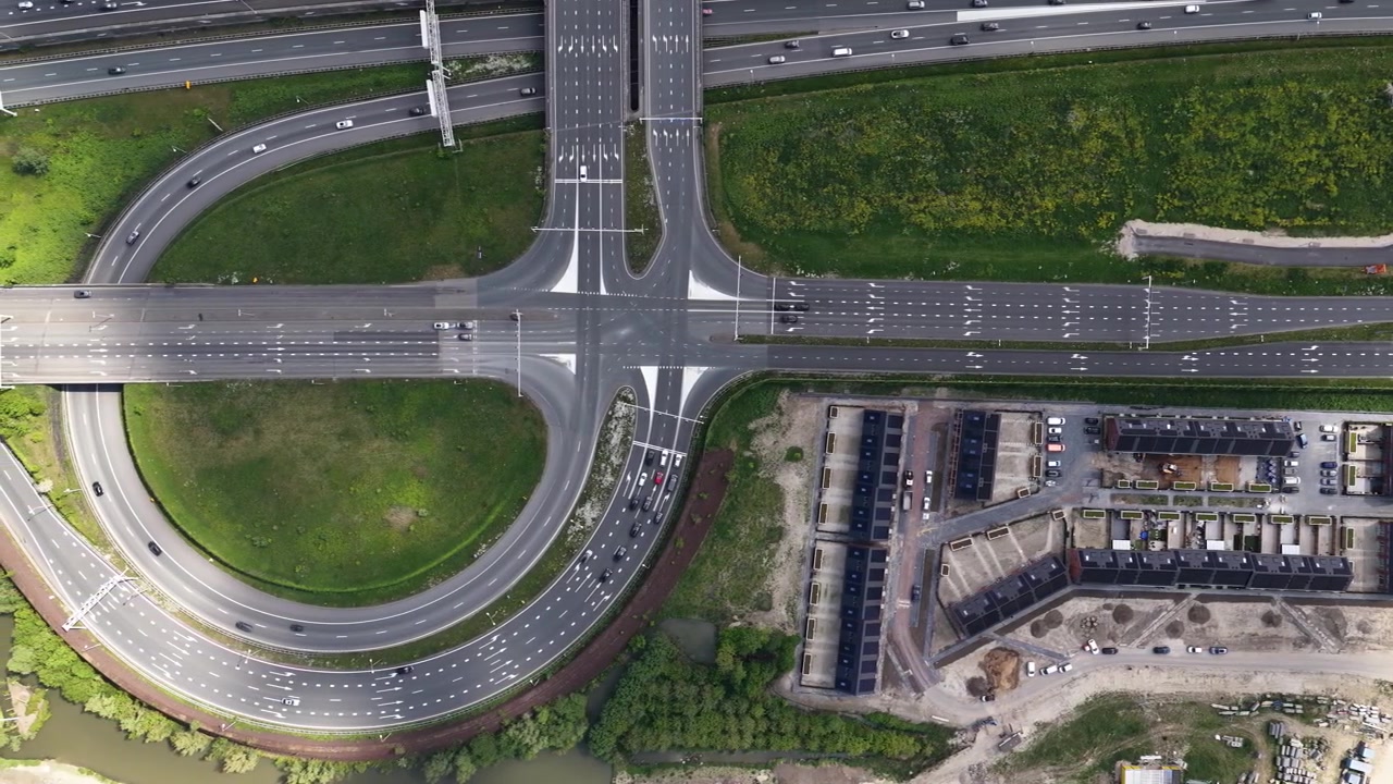 Dynamic aerial perspective showing bustling traffic flow on a complex highway interchange next to a new suburban residential neighborhood development with modern houses under construction