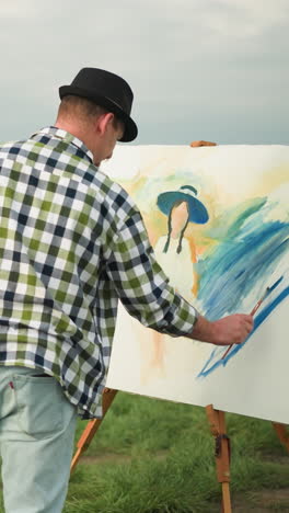 back view of an artist wearing a black hat and checkered shirt, holding a palette while painting a large, blue-themed canvas in a peaceful grass field