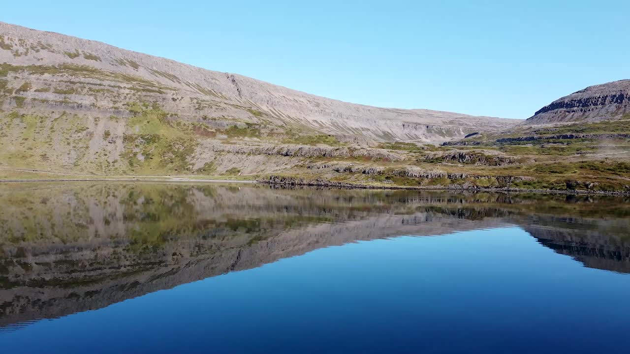 Drone view of calm fjord with mirror-like water perfectly reflecting rugged cliffs and clear blue sky, showcasing peaceful natural beauty and untouched scenery of remote Icelandic wilderness