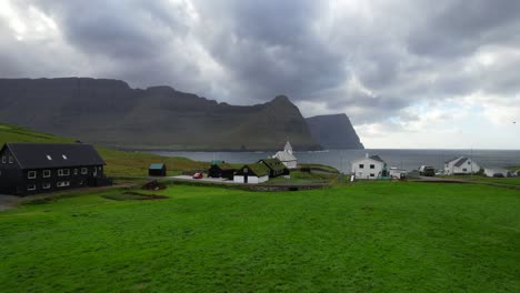 Aerial-through-a-meadow-over-traditional-Vidareidi-village,-turf-roofs-and-old-stone-church