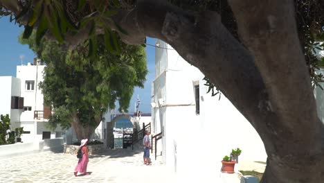 Reveal-behind-trees-of-elder-tourist-couple-in-front-of-typical-white,-Greek-church-with-cobbled-ground-SLOW-MOTION