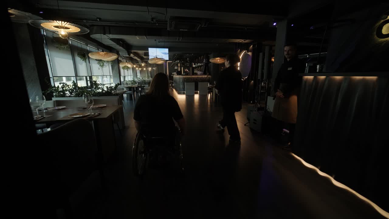 체어에 앉은 한 여성이 현대적인 장식과 예술적인 요소를 가진 세련되고 희미하게 조명된 레스토랑에 들어갑니다.이 장면은 유행의 식사 환경에서 접근성을 강조하며 이동성과 포적 인 디자인을 강조합니다.