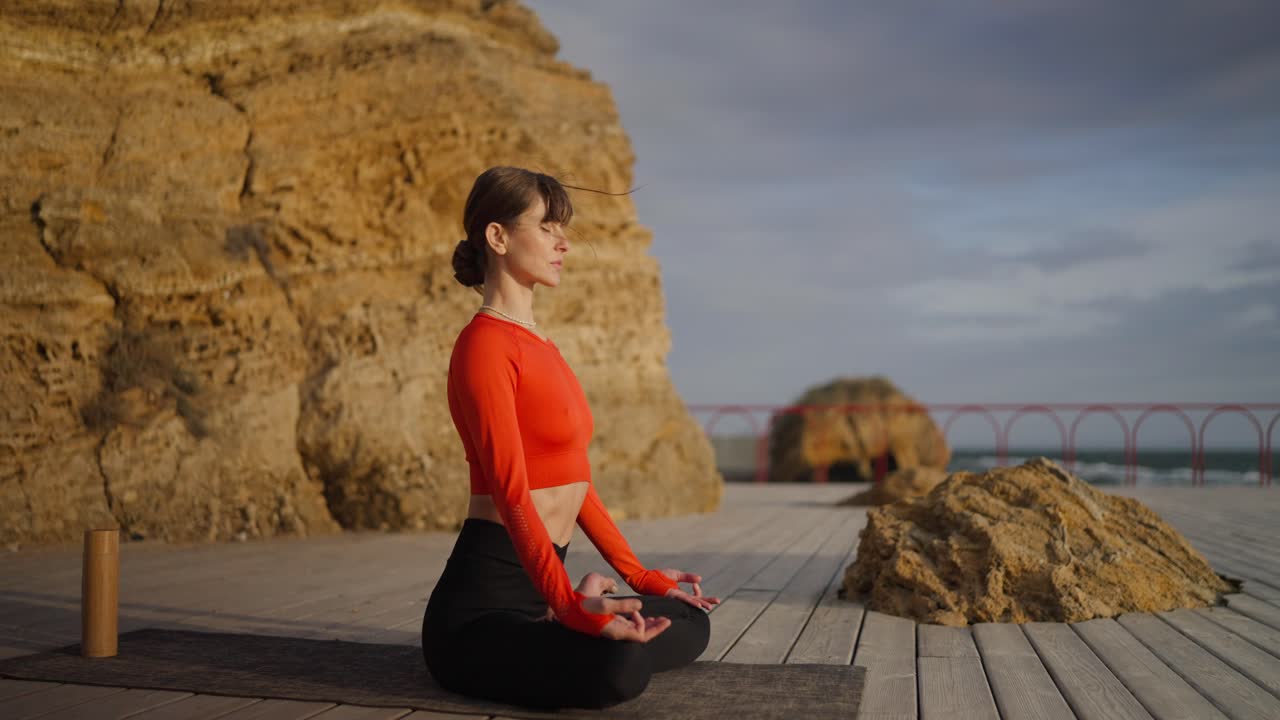 una mujer meditando en una cubierta de madera con vistas al océano durante la puesta de sol.