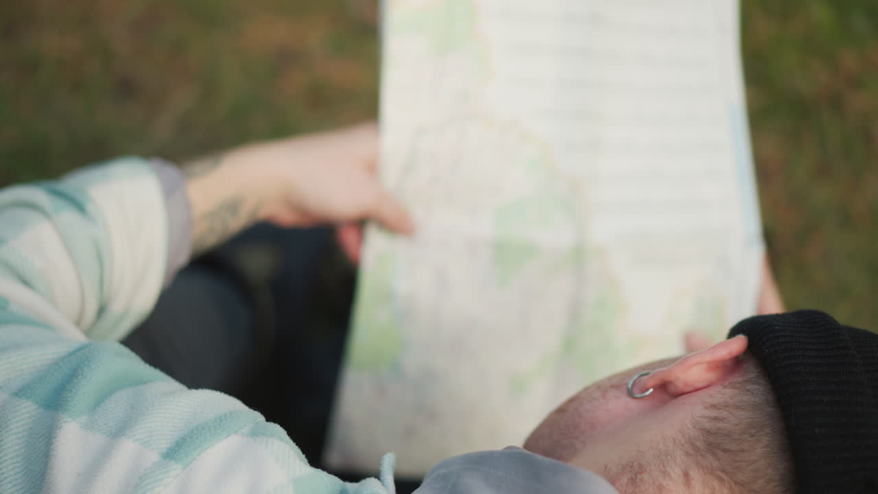 white male backpacker studying map on lap in meadow, reflective pause between hikes, knees and jacket visible, tracing waypoints and planning next stage, close aerial perspective with soft light