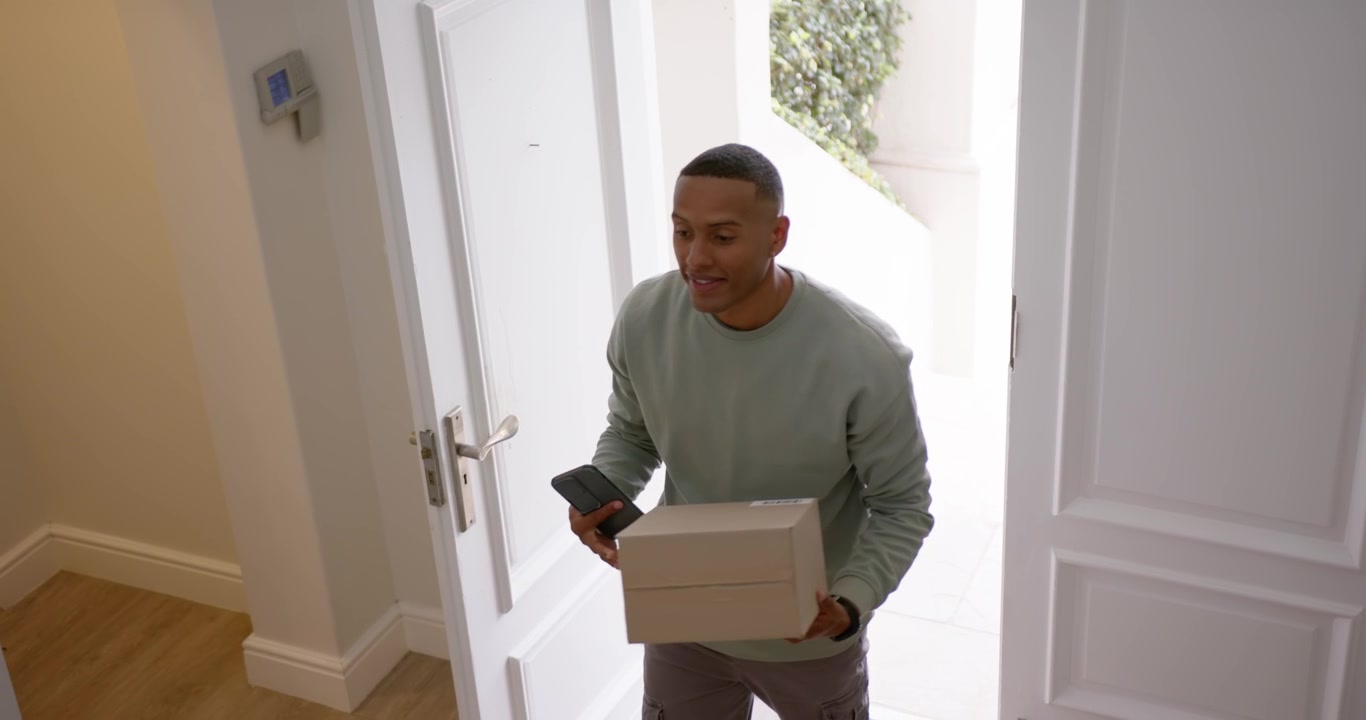 African American adult male checking phone while carrying box, stepping through front doorway