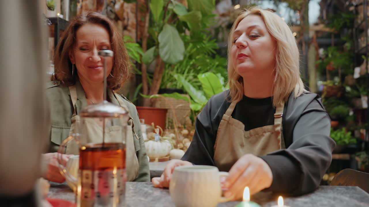 Due donne sorseggiano tè a un tavolo per il rinvaso in un negozio di fiori, ridendo e chiacchierando tra piante rigogliose, cesti di vimini, tazze di ceramica, una teiera di vetro, candele, grembiuli e un'illuminazione calda, mentre le mani sistemano gli steli