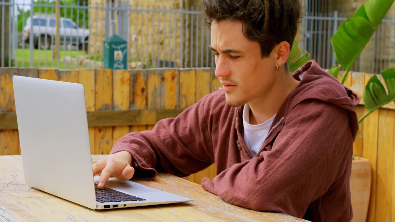 vista lateral de un joven skateboarder caucásico trabajando en una computadora portátil en un café al aire libre 4k