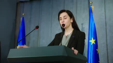 Female-politician-makes-an-announcement,-answers-journalists-questions-and-gives-interview-for-media.-Confident-representative-of-the-European-Union-during-press-conference.-Backdrop-with-EU-flags.