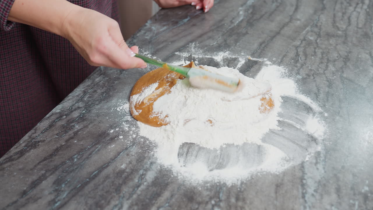 primer plano de una mujer usando una espátula verde para mezclar masa marrón gruesa con harina en el mostrador, preparación de horneado en la cocina, mezcla de ingredientes para pasteles caseros