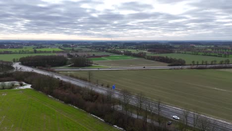 sunny and cloudy winter day in german rural town. farm fields with driving cars on intersection and autobahn highway. aerial forward wide shot. leafless trees in idyllic scenery.