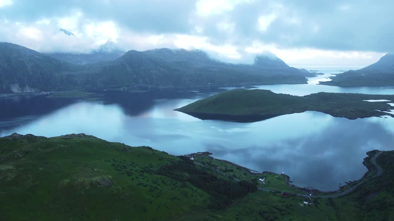 Aerial drone fotoage showing large Lofoten Norway green mossy and wet mountains that are surrounding a large lake or blue river during a cloudy and misty day. Large blue and white clouds over mountain
