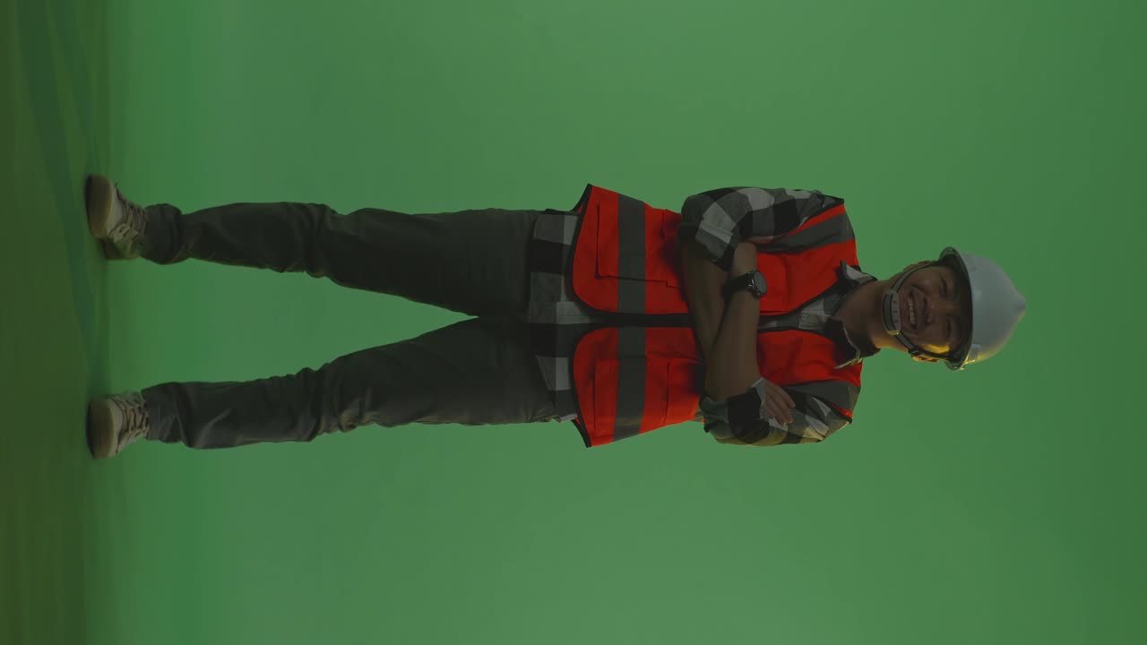 cuerpo lleno de ingeniero masculino asiático con casco de seguridad cruzando los brazos y sonriendo a la cámara mientras está de pie en el estudio de fondo de pantalla verde, durante la puesta o el amanecer