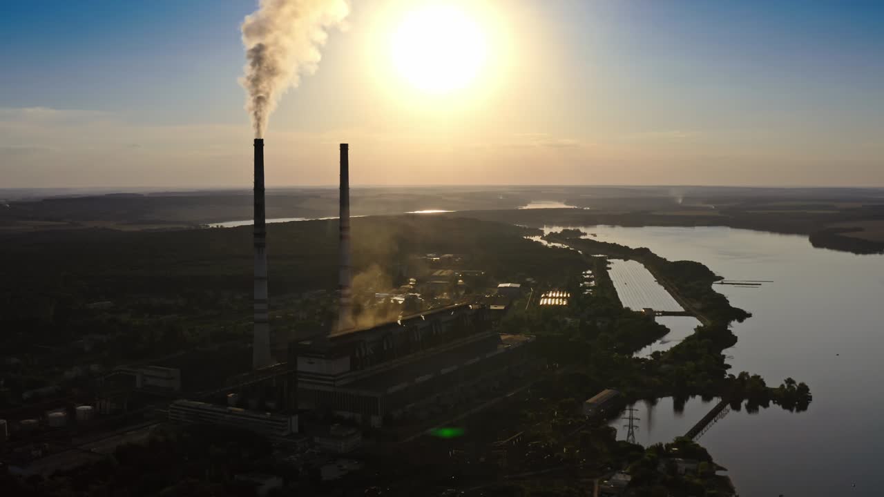 Fabbrica scura contro il sole arancione al tramonto. Ciminiere della fabbrica con fumo. Fumo sporco fuoriesce dalle ciminiere nell'aria sullo sfondo del fiume. Video con vista aerea