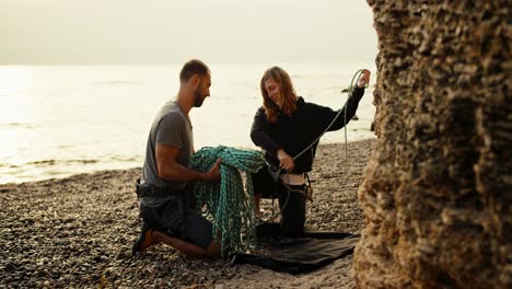 A-blonde-girl-with-curly-hair-in-black-clothes-untangles-and-unties-a-special-rope-together-with-her-boyfriend-in-order-to-start-rock-climbing-on-a-rocky-shore-near-the-rocks