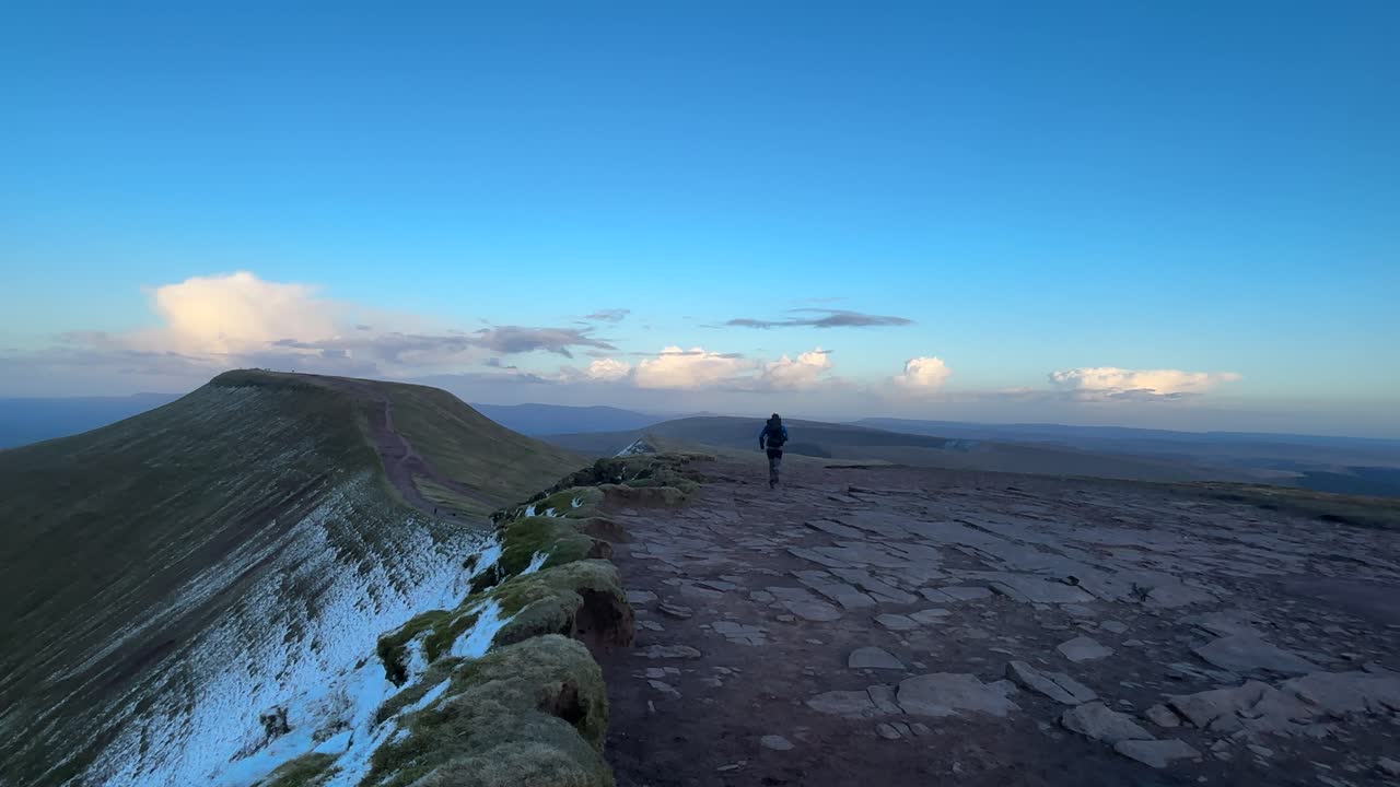 Escursionista che corre lungo la cresta sommitale vicino a Pen y Fan in Galles, Regno Unito, con terreno roccioso, ampio orizzonte e cielo blu che creano un suggestivo paesaggio per avventure all'aperto.