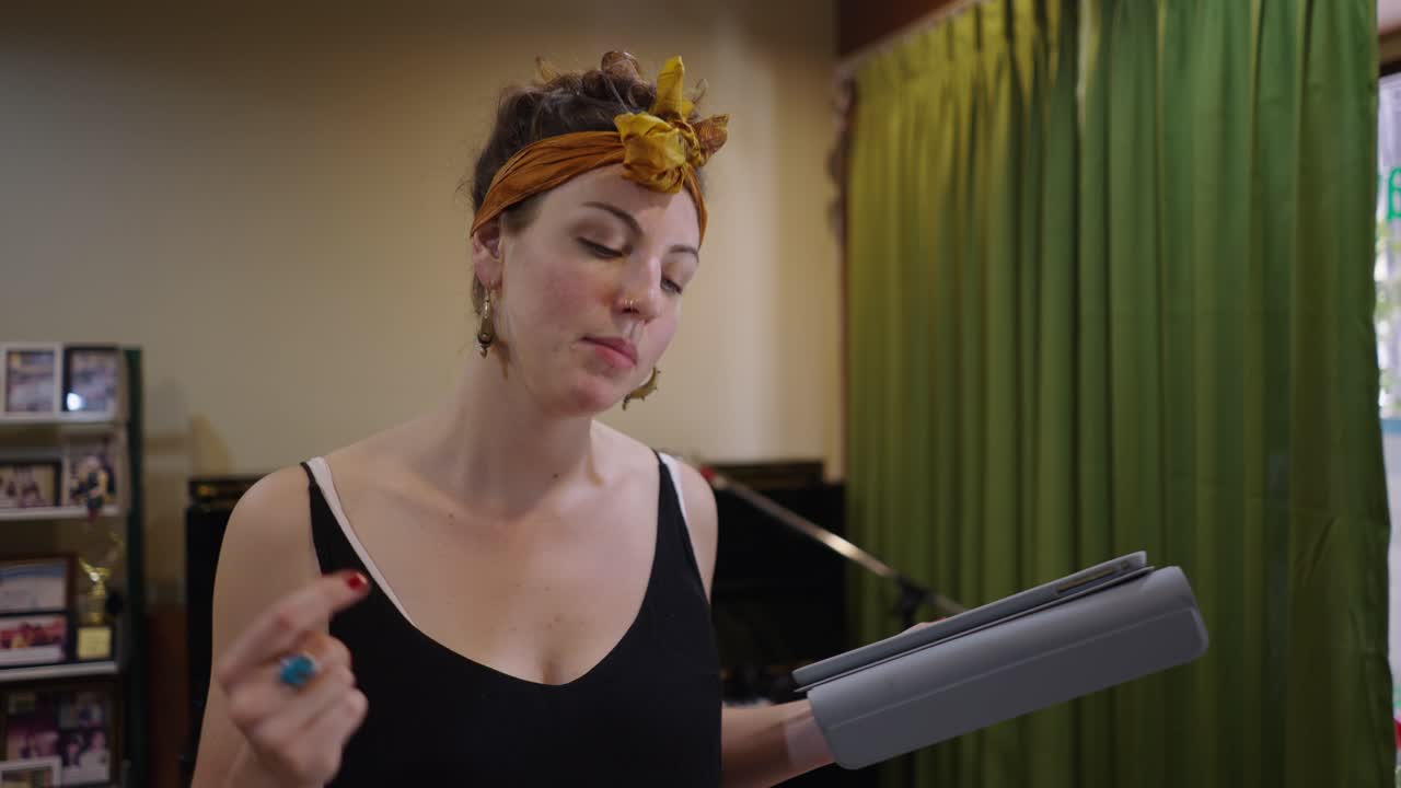 A woman holds a tablet in an indoor setting with a piano and green curtains