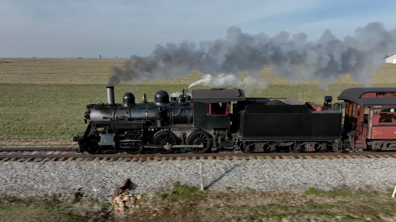 A vintage steam train moves along the tracks, emitting smoke into the blue sky while crossing a vast, green agricultural landscape during the daytime