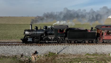 a vintage steam train moves along the tracks, emitting smoke into the blue sky while crossing a vast, green agricultural landscape during the daytime