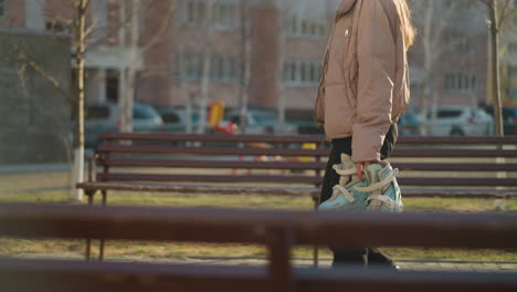 a person dressed in a peach jacket and black trousers walks through a sunlit park, holding a pair of rollerblades in one hand. benches and residential buildings are visible in the background