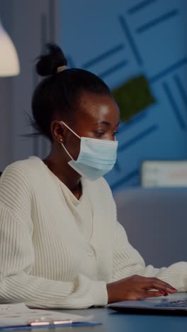 vertical video african employee with protection face mask working at laptop overtime
