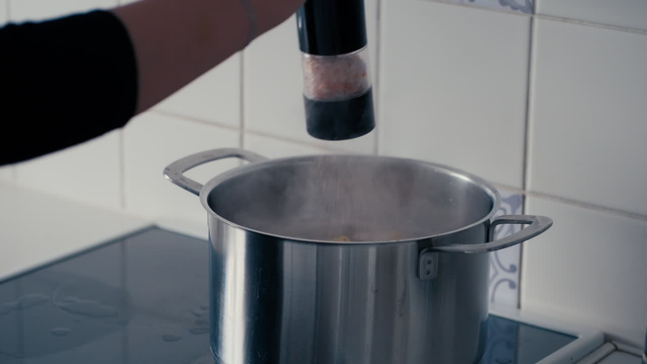 Woman's Hand opening the Lid of a big steel Pot and adding Himalayan Salt to boiling Water with Pasta in slow motion