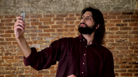 joven atractivo hombre moreno con barba está haciendo video en el teléfono inteligente, tonto, riendo, fondo de ladrillo, concepto de comunicación