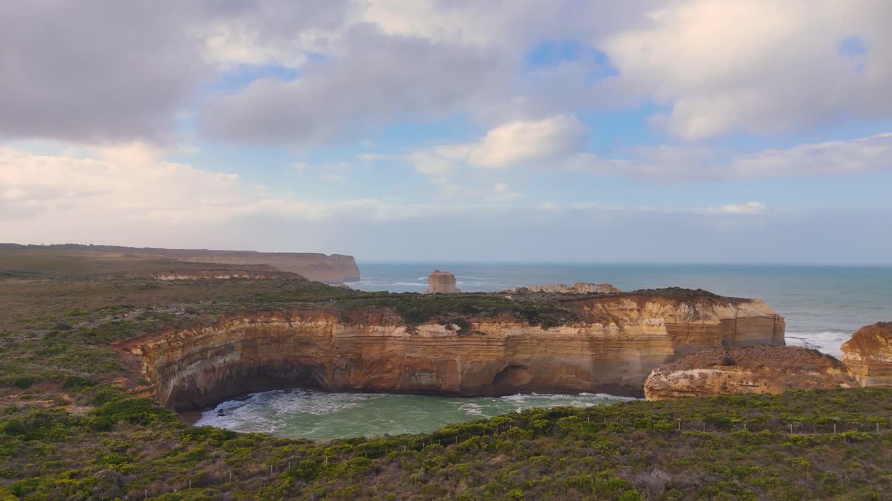 Drone footage captures the rugged cliffs and ocean waves at Loch Ard Gorge, Port Campbell, under a partly cloudy sky