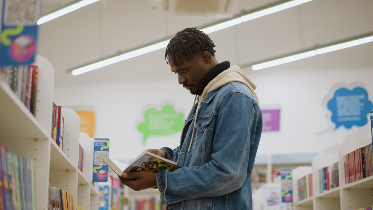Big boy reading book in library. Focused young man in denim jacket holding book, carefully inspecting pages while surrounded by organized shelves with colorful books in modern library setting