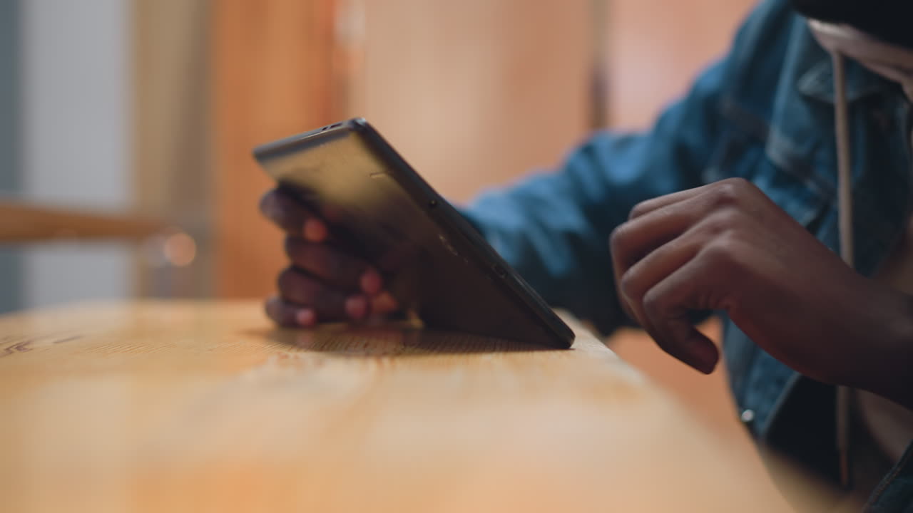 close up of black hand holding tablet at angle on wooden table while other hand taps screen indoors with denim sleeve blurred warm bokeh background conveying modern digital leisure moment and relaxation