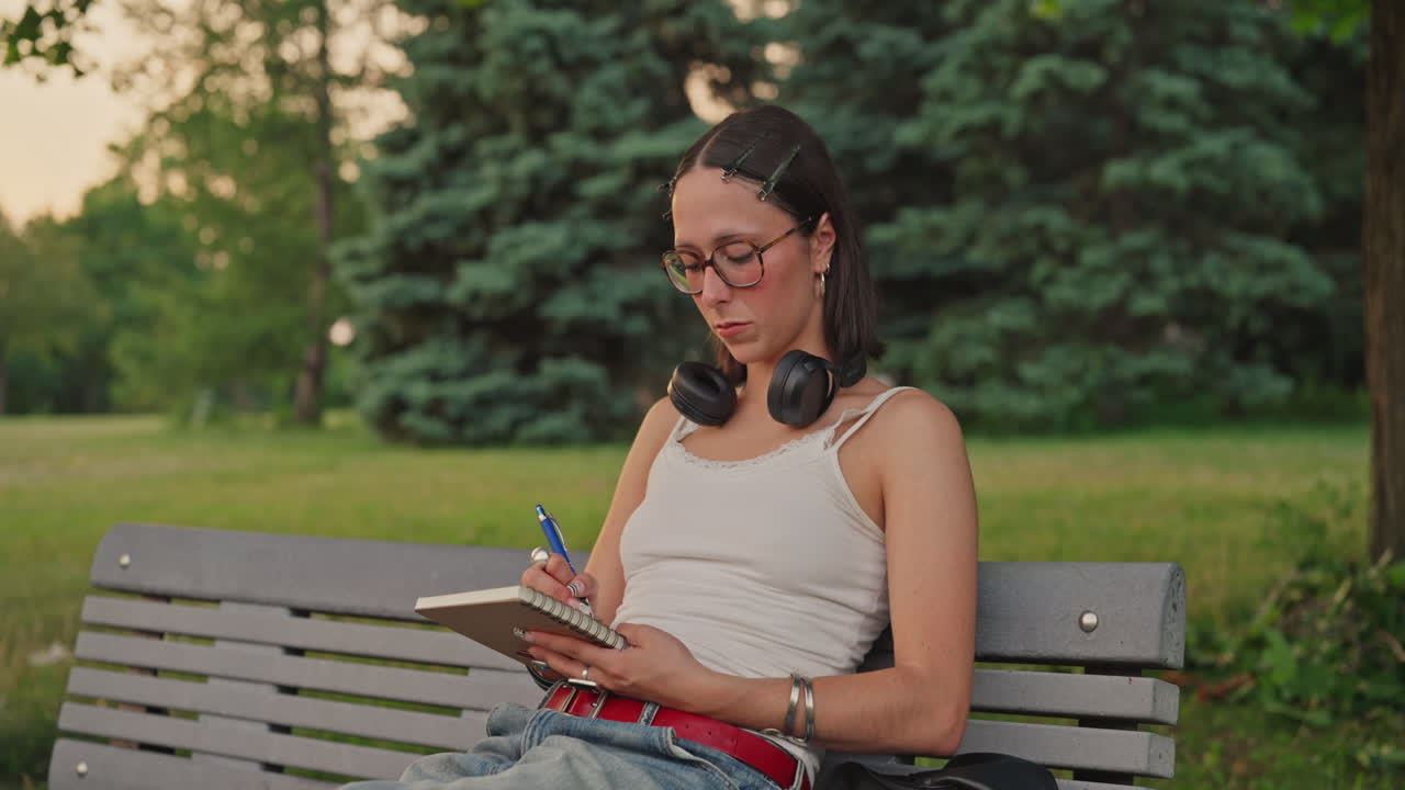 Young woman journaling on park bench, headphones around neck, reflective solitude moment