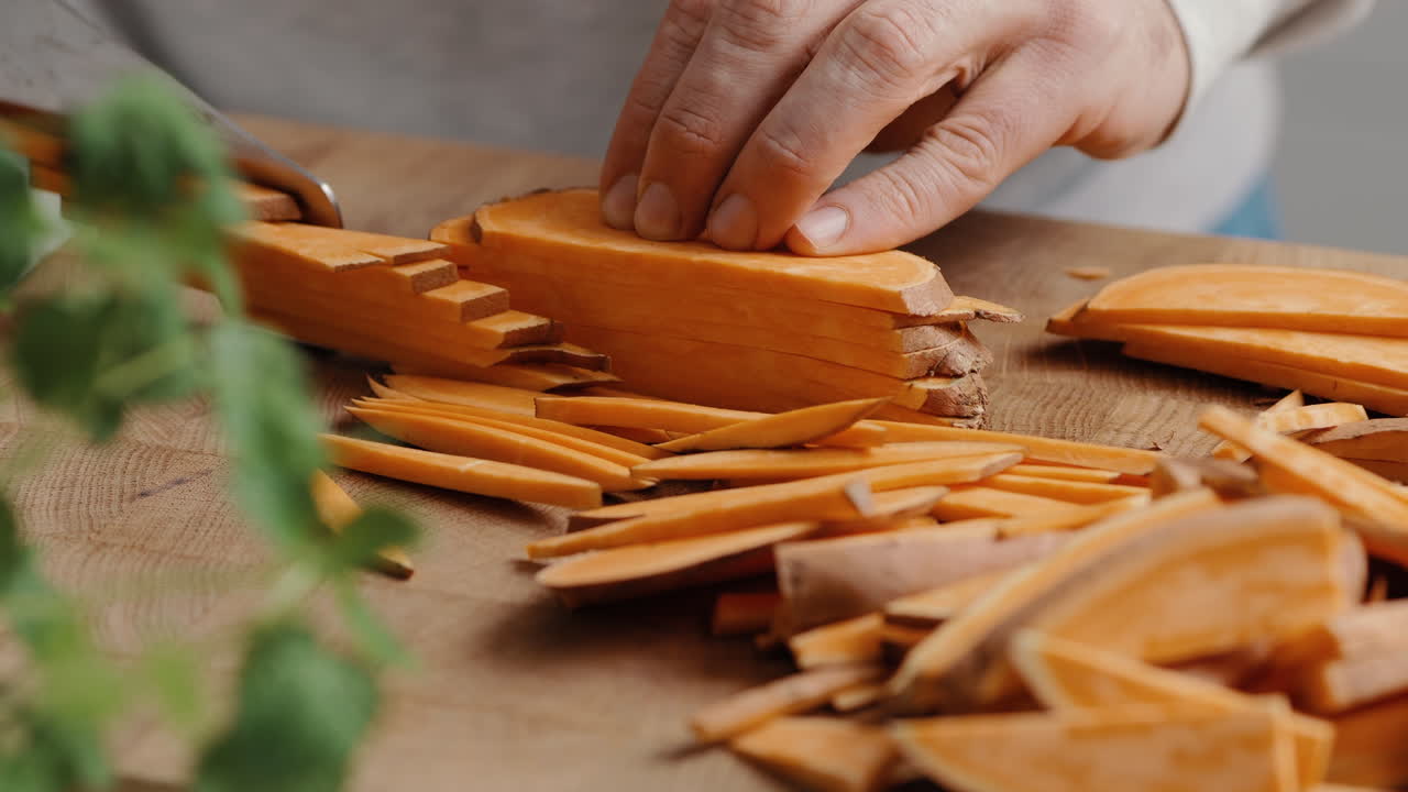Male chef sliceing sweet potato in thin slices for sweet potato fries in the kitchen on a wooden cutting board in the kitchen