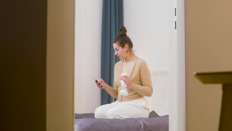 Young-Mother-Using-Breast-Pump-And-Scrolling-On-Mobile-Phone-While-Sitting-On-The-Bed-At-Home