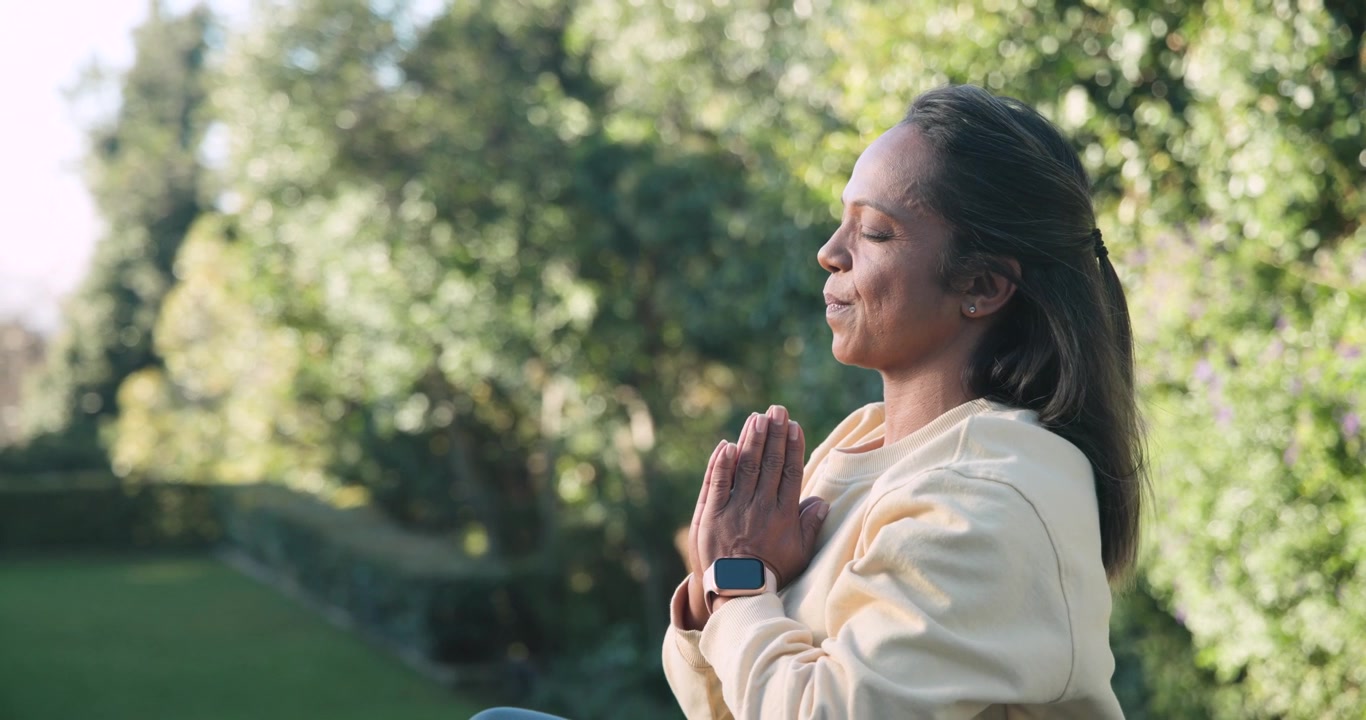 Mature African woman breathing, stretching into prayer in garden, sweatshirt smartwatch, copy space