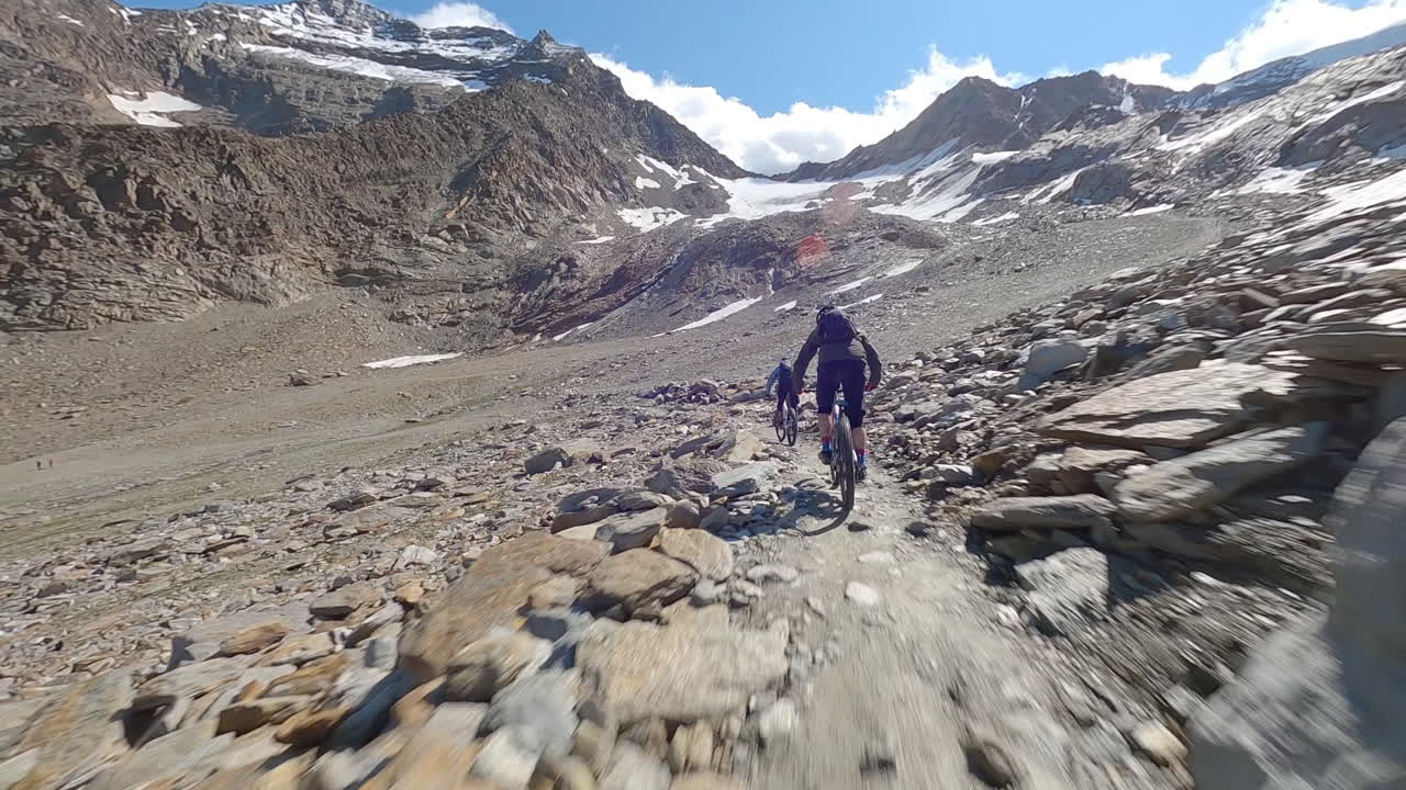 FPV drone follows two mountain bikers descending steep, exposed alpine trails above Saas-Grund and Saas-Fee on a clear bluebird summer day, showcasing epic terrain and speed