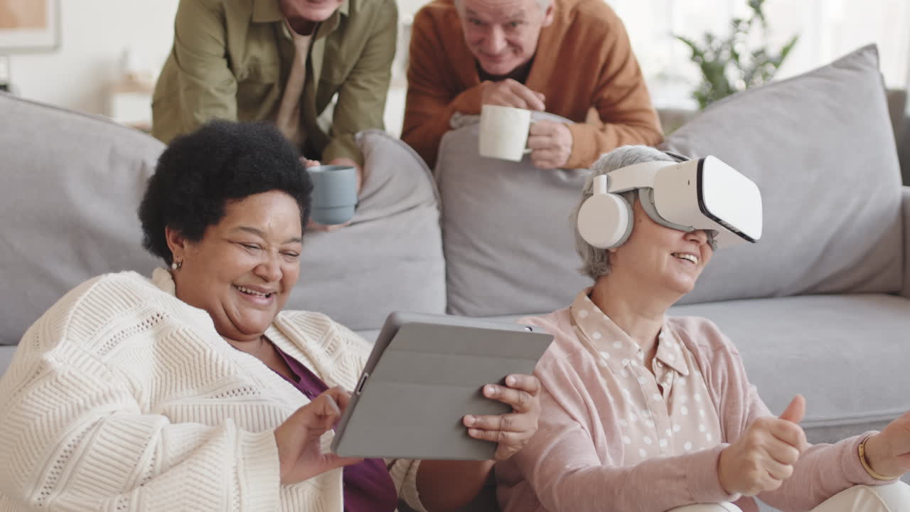 Medium close-up of diverse senior female friends leaning on couch, grey-haired woman using VR headset, African lady having tablet computer, laughing, two men holding cups looking at them