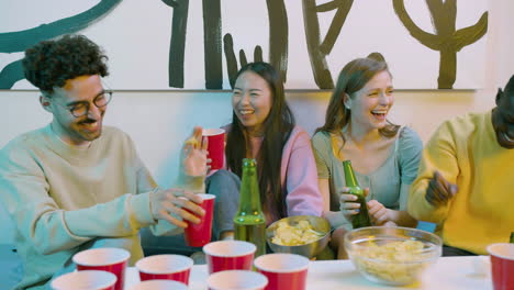 felices jóvenes amigos multiétnicos bebiendo cerveza, comiendo papas fritas y riendo mientras se sientan en el sofá en casa